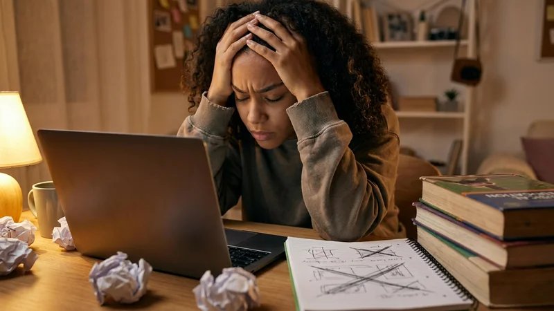 Estudante com expressão frustrada diante do computador, papéis amassados ao redor, representando dificuldades com TCC